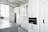 A modern kitchen features sleek white cabinetry, a built-in oven, and a large countertop island with a stovetop. The space is brightly lit by a large window, and a vase with green foliage sits on the counter. Stainless steel appliances add to the contemporary feel.