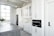 A modern kitchen features sleek white cabinetry, a built-in oven, and a large countertop island with a stovetop. The space is brightly lit by a large window, and a vase with green foliage sits on the counter. Stainless steel appliances add to the contemporary feel.
