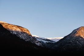 A majestic landscape featuring two mountain peaks with their tops illuminated by the warm glow of sunlight. Snow covers parts of the peaks, contrasted by a deep blue sky. The mountains create a sense of depth and tranquility.