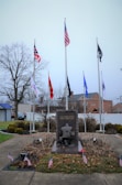 A memorial monument with a soldier kneeling, surrounded by multiple flags, including the American flag, and small American flags around its base. The background includes a few trees and buildings under an overcast sky. The monument has the inscription 'ALL GAVE SOME, SOME GAVE ALL.'