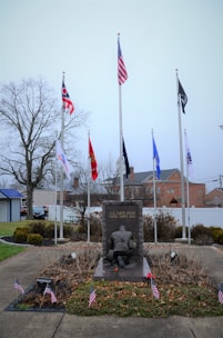 A memorial monument with a soldier kneeling, surrounded by multiple flags, including the American flag, and small American flags around its base. The background includes a few trees and buildings under an overcast sky. The monument has the inscription 'ALL GAVE SOME, SOME GAVE ALL.'