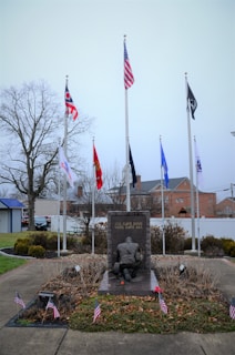 A memorial monument with a soldier kneeling, surrounded by multiple flags, including the American flag, and small American flags around its base. The background includes a few trees and buildings under an overcast sky. The monument has the inscription 'ALL GAVE SOME, SOME GAVE ALL.'