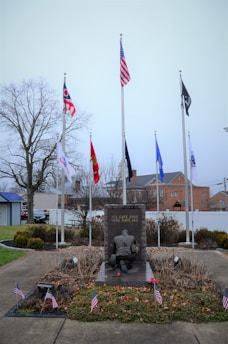 A memorial monument with a soldier kneeling, surrounded by multiple flags, including the American flag, and small American flags around its base. The background includes a few trees and buildings under an overcast sky. The monument has the inscription 'ALL GAVE SOME, SOME GAVE ALL.'