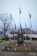 A memorial monument with a soldier kneeling, surrounded by multiple flags, including the American flag, and small American flags around its base. The background includes a few trees and buildings under an overcast sky. The monument has the inscription 'ALL GAVE SOME, SOME GAVE ALL.'