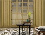 A stylish interior setting features a large wooden window with multiple panes, allowing soft natural light to filter in. The foreground is adorned with two wooden tables; one holds a decorative plant arrangement in a glass vase, while the other supports a stack of books and magazines. The floor is patterned with geometric tiles, adding to the room's elegant ambiance.