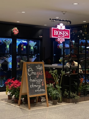 A flower shop with a neon sign reading 'Roses are red', featuring a variety of plants and flower arrangements on display. A chalkboard with a handwritten message is placed at the entrance. The interior includes lighted shelves with assorted bouquets.
