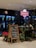 A flower shop with a neon sign reading 'Roses are red', featuring a variety of plants and flower arrangements on display. A chalkboard with a handwritten message is placed at the entrance. The interior includes lighted shelves with assorted bouquets.