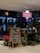 A flower shop with a neon sign reading 'Roses are red', featuring a variety of plants and flower arrangements on display. A chalkboard with a handwritten message is placed at the entrance. The interior includes lighted shelves with assorted bouquets.