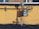 A gas meter is mounted on a yellow wall with visible pipes running horizontally and vertically. The shadows of the pipes and meter are cast sharply on the wall. The ground below the wall is bare with a few sparse plants.