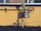 A gas meter is mounted on a yellow wall with visible pipes running horizontally and vertically. The shadows of the pipes and meter are cast sharply on the wall. The ground below the wall is bare with a few sparse plants.