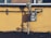A gas meter is mounted on a yellow wall with visible pipes running horizontally and vertically. The shadows of the pipes and meter are cast sharply on the wall. The ground below the wall is bare with a few sparse plants.