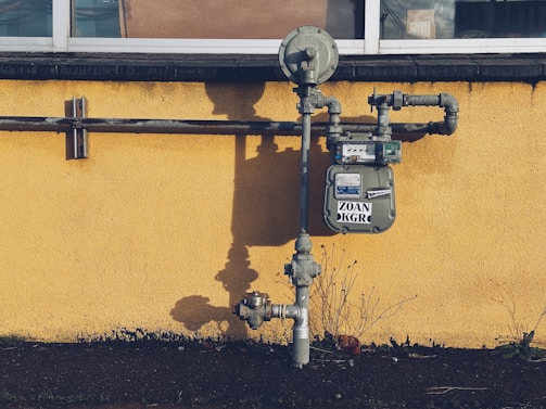 A gas meter is mounted on a yellow wall with visible pipes running horizontally and vertically. The shadows of the pipes and meter are cast sharply on the wall. The ground below the wall is bare with a few sparse plants.