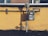 A gas meter is mounted on a yellow wall with visible pipes running horizontally and vertically. The shadows of the pipes and meter are cast sharply on the wall. The ground below the wall is bare with a few sparse plants.