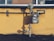 A gas meter is mounted on a yellow wall with visible pipes running horizontally and vertically. The shadows of the pipes and meter are cast sharply on the wall. The ground below the wall is bare with a few sparse plants.