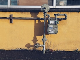 A gas meter is mounted on a yellow wall with visible pipes running horizontally and vertically. The shadows of the pipes and meter are cast sharply on the wall. The ground below the wall is bare with a few sparse plants.
