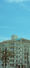 A mid-rise, modern apartment building with multiple balconies is set against a bright blue sky. The building is equipped with numerous air conditioning units, and there are some trees in the foreground. Utility poles and cables are also visible, adding to the urban environment.