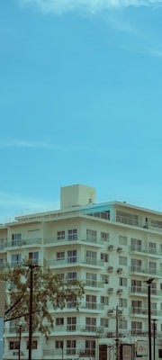 A mid-rise, modern apartment building with multiple balconies is set against a bright blue sky. The building is equipped with numerous air conditioning units, and there are some trees in the foreground. Utility poles and cables are also visible, adding to the urban environment.