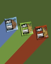 Three packs of energy cookies are displayed against a background with diagonal color stripes. The packs are in different colors: blue, green, and orange, each with distinct labeling and branding.
