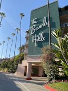 A large, stylish building with a prominent sign reading 'The Beverly Hills'. Tall palm trees line the entrance, surrounded by lush green foliage with some bright red flowers. The building exterior is painted green with decorative railings.