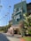 A large, stylish building with a prominent sign reading 'The Beverly Hills'. Tall palm trees line the entrance, surrounded by lush green foliage with some bright red flowers. The building exterior is painted green with decorative railings.