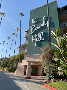 A large, stylish building with a prominent sign reading 'The Beverly Hills'. Tall palm trees line the entrance, surrounded by lush green foliage with some bright red flowers. The building exterior is painted green with decorative railings.