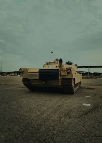 A military tank is positioned on an open, paved area. The tank is tan-colored and has various equipment mounted on top. The sky is overcast and there is no other significant activity around. Some small buildings and structures are visible in the distant background.