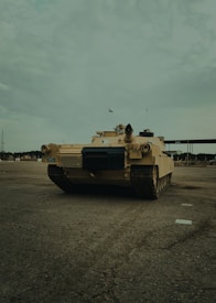 A military tank is positioned on an open, paved area. The tank is tan-colored and has various equipment mounted on top. The sky is overcast and there is no other significant activity around. Some small buildings and structures are visible in the distant background.