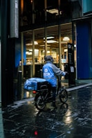A delivery person wearing a blue jacket and helmet is sitting on a bicycle on a wet street next to modern office buildings with large glass windows. The jacket and delivery bag have logos and text on them.