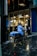 A delivery person wearing a blue jacket and helmet is sitting on a bicycle on a wet street next to modern office buildings with large glass windows. The jacket and delivery bag have logos and text on them.
