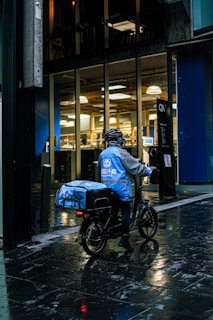 A delivery person wearing a blue jacket and helmet is sitting on a bicycle on a wet street next to modern office buildings with large glass windows. The jacket and delivery bag have logos and text on them.