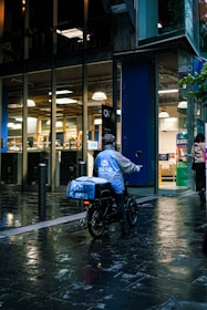 A food delivery person on an electric bike is riding in a wet urban street next to a modern building with large glass windows. The delivery person is wearing a blue jacket and helmet, and the evening lighting casts reflections on the wet pavement.