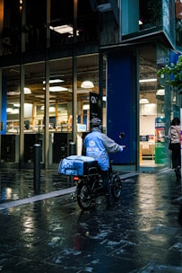 A food delivery person on an electric bike is riding in a wet urban street next to a modern building with large glass windows. The delivery person is wearing a blue jacket and helmet, and the evening lighting casts reflections on the wet pavement.