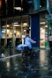A food delivery person on an electric bike is riding in a wet urban street next to a modern building with large glass windows. The delivery person is wearing a blue jacket and helmet, and the evening lighting casts reflections on the wet pavement.