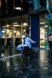 A food delivery person on an electric bike is riding in a wet urban street next to a modern building with large glass windows. The delivery person is wearing a blue jacket and helmet, and the evening lighting casts reflections on the wet pavement.