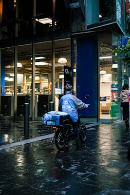 A food delivery person on an electric bike is riding in a wet urban street next to a modern building with large glass windows. The delivery person is wearing a blue jacket and helmet, and the evening lighting casts reflections on the wet pavement.
