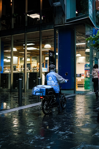 A food delivery person on an electric bike is riding in a wet urban street next to a modern building with large glass windows. The delivery person is wearing a blue jacket and helmet, and the evening lighting casts reflections on the wet pavement.