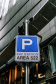 A blue parking sign indicates a pay station for area 522, mounted on a metal pole. There is a security camera attached above the sign. The background consists of modern architectural structures with glass and metal elements.