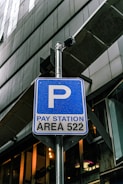 A blue parking sign indicates a pay station for area 522, mounted on a metal pole. There is a security camera attached above the sign. The background consists of modern architectural structures with glass and metal elements.