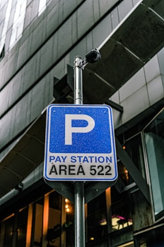 A blue parking sign indicates a pay station for area 522, mounted on a metal pole. There is a security camera attached above the sign. The background consists of modern architectural structures with glass and metal elements.