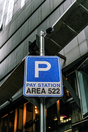 A blue parking sign indicates a pay station for area 522, mounted on a metal pole. There is a security camera attached above the sign. The background consists of modern architectural structures with glass and metal elements.