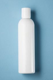A plain white plastic bottle with a cylindrical shape and a cap on top, positioned vertically against a light blue background.