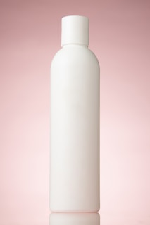A plain, white cylindrical bottle with a pink background. The bottle has a rounded cap and no labels or markings, reflecting a minimalist design.