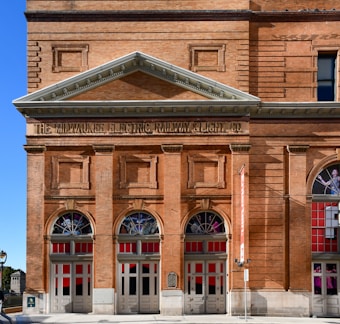 A historical brick building with classical architecture featuring three large arched windows. The building facade displays the inscription 'THE MILWAUKEE ELECTRIC RAILWAY & LIGHT CO.' in bold letters above the entrance. Below the arches, there are red and white doors. A banner hangs on the right side of the facade, and a plaque is visible near the middle door.