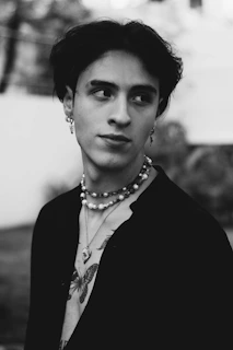 A black and white portrait of a person with short dark hair, wearing multiple necklaces and earrings. The person has a tattoo of a butterfly and appears to be looking off to the side, with a neutral to contemplative expression.