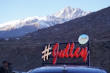 A scenic view of snow-capped mountains in the background with a hilly terrain in the foreground. A bright sign with the hashtag '#Julley' is prominently displayed, associated with the 'Lahaul Administration'. A few people are visible near a car at the bottom of the image.