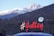 A scenic view of snow-capped mountains in the background with a hilly terrain in the foreground. A bright sign with the hashtag '#Julley' is prominently displayed, associated with the 'Lahaul Administration'. A few people are visible near a car at the bottom of the image.