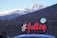 A scenic view of snow-capped mountains in the background with a hilly terrain in the foreground. A bright sign with the hashtag '#Julley' is prominently displayed, associated with the 'Lahaul Administration'. A few people are visible near a car at the bottom of the image.