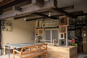 A rustic outdoor setting featuring a wooden table and bench beneath an overhang with exposed wooden beams. The wall is adorned with a collection of variously framed windows and an assortment of retro items, including a colorful ride-on toy horse in the background. The area has a mix of modern and vintage elements, with utility boxes mounted on the wall.