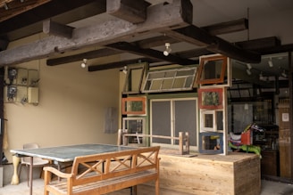 A rustic outdoor setting featuring a wooden table and bench beneath an overhang with exposed wooden beams. The wall is adorned with a collection of variously framed windows and an assortment of retro items, including a colorful ride-on toy horse in the background. The area has a mix of modern and vintage elements, with utility boxes mounted on the wall.