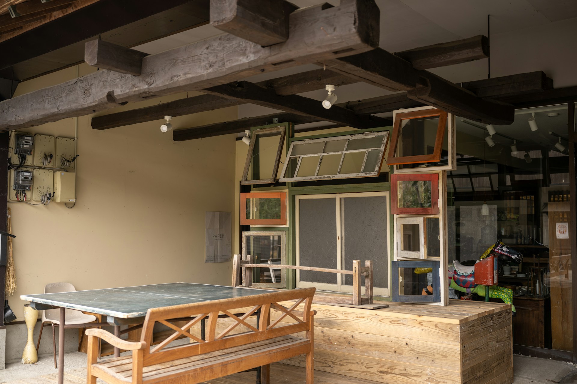A rustic outdoor setting featuring a wooden table and bench beneath an overhang with exposed wooden beams. The wall is adorned with a collection of variously framed windows and an assortment of retro items, including a colorful ride-on toy horse in the background. The area has a mix of modern and vintage elements, with utility boxes mounted on the wall.
